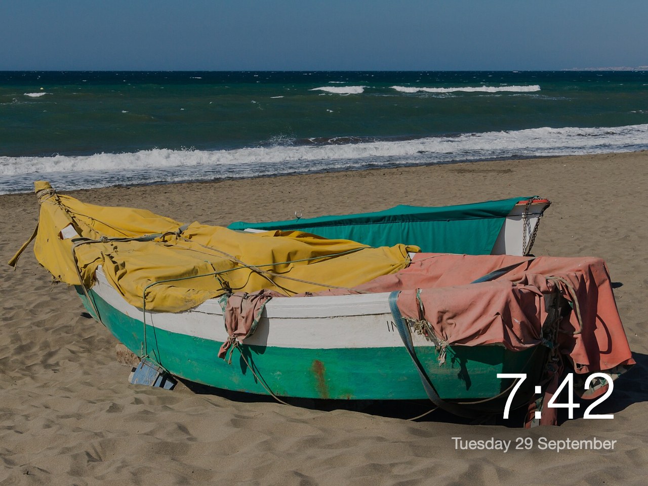 A photo of fishing boats on a beach filling the screen, with the time and date in the corner.